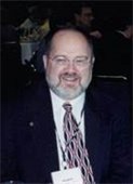 Smiling man with glasses, beard, and dark suit seated at an event. He wears a patterned tie and name tag, surrounded by a dimly lit background.