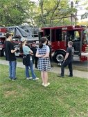 teen participants watch as a firefighter dressed in firefighting gear talks in front of a fire truck parked streetside