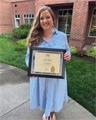 woman wearing a blue and white striped dress smiling and holding her diploma outside of a building on a sunny day