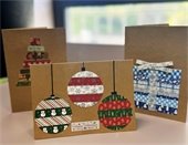 Three festive holiday cards are displayed. One shows ornaments, another a Christmas tree, and the last a gift box with a bow, all on brown kraft paper.
