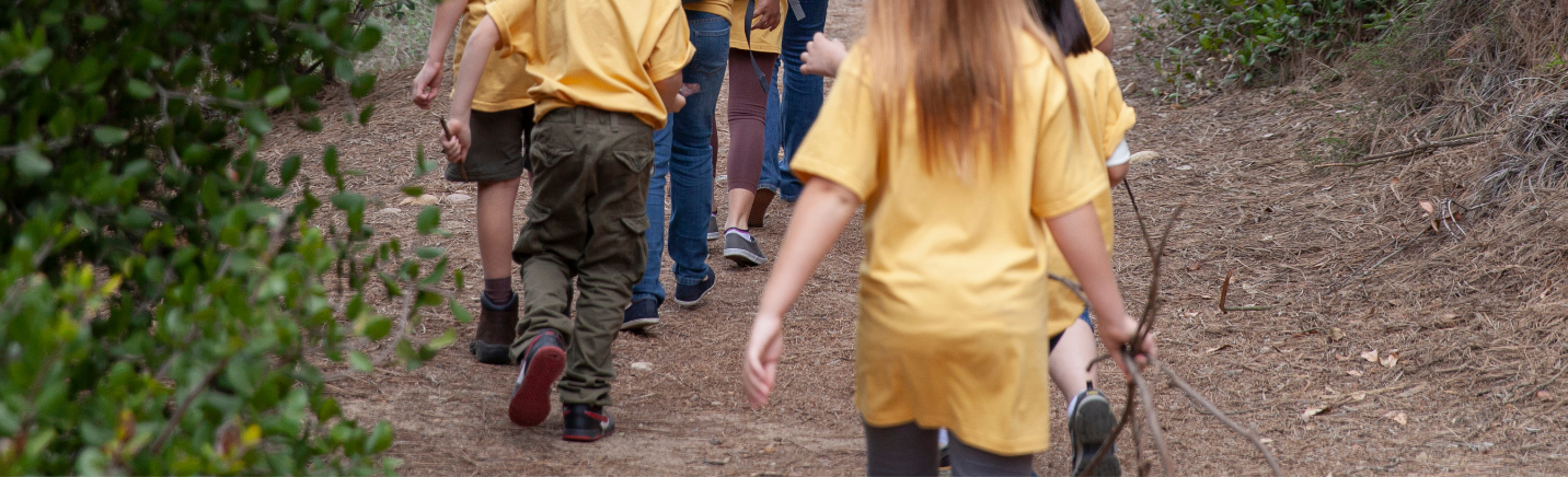 Group of children walking along a dirt trail in a wooded area, wearing yellow shirts.