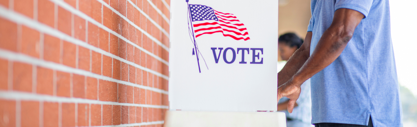 Person places a ballot into a white voting box with an American flag and the word VOTE.